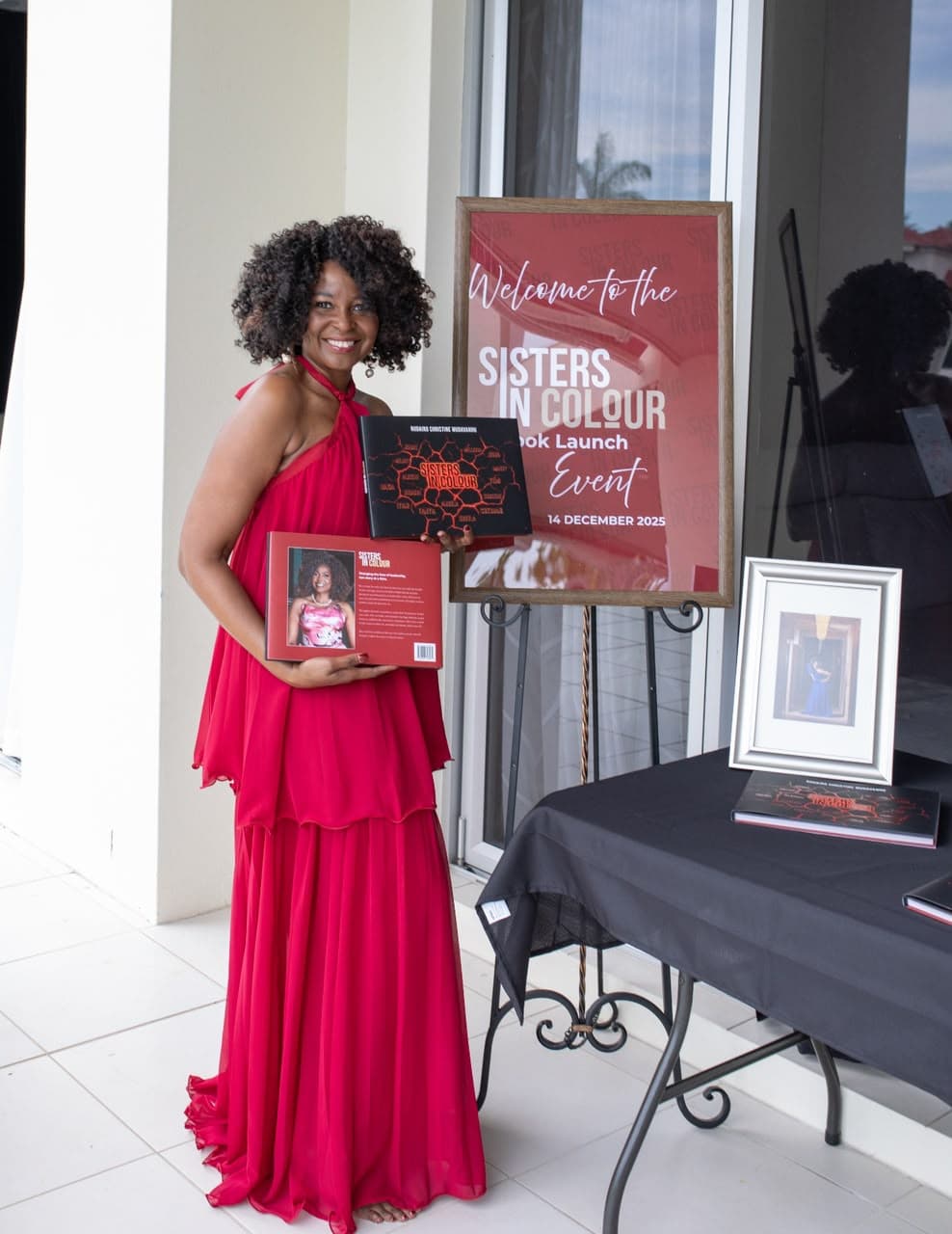 Woman holding Sisters in Colour books beside the Book Launch welcome sign